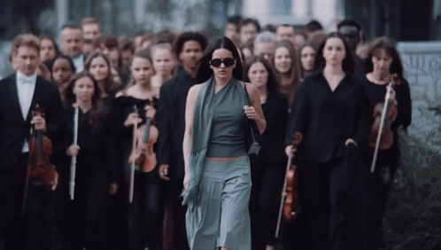 A Woman Wearing Sunglasses Walks In Front Of A Large Group Of Musicians GIF