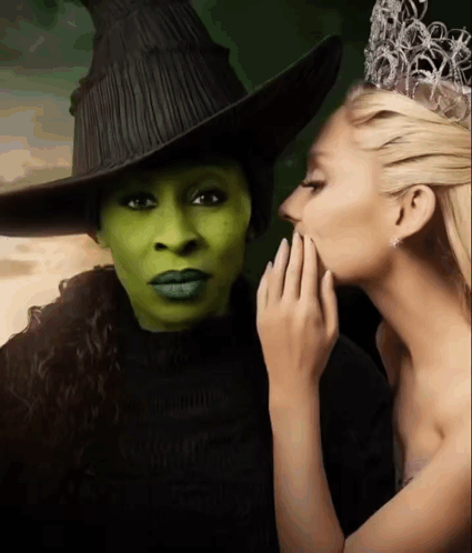 A Woman Whispering Into Another Woman 'S Ear While Wearing A Witch Costume GIF