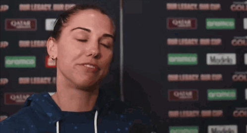 A Woman Wipes Her Face With A Towel In Front Of A Wall That Says Qatar On It GIF