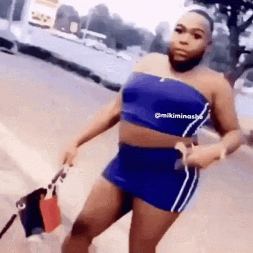 A Woman With A Beard Is Wearing A Blue Top And Shorts And Holding A Bag . GIF
