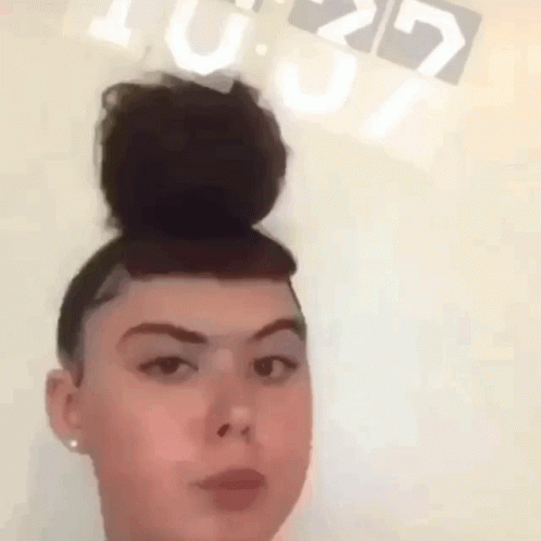 A Woman With A Bun On Her Head Is Standing In Front Of A Clock That Reads 10:29 GIF