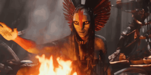 A Woman With A Feathered Headdress Is Standing In Front Of A Fire GIF