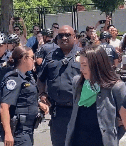 A Woman With A Green Bandana On Her Neck Is Being Escorted By Police GIF