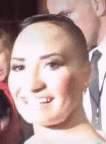 A Woman With A Shaved Head Is Smiling In Front Of A Man In A Suit And Tie . GIF