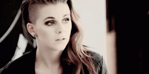 A Woman With A Shaved Head Is Wearing Earrings And A Black Jacket . GIF