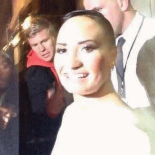 A Woman With A Shaved Head Smiles In Front Of A Man In A Suit GIF