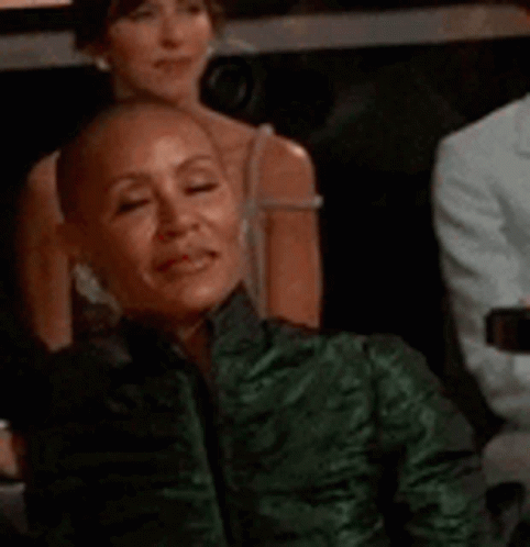 A Woman With A Shaved Head Wearing A Green Jacket Is Sitting In A Theatre . GIF
