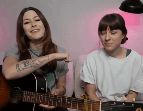 A Woman With A Tattoo On Her Arm Is Playing A Guitar Next To Another Woman GIF