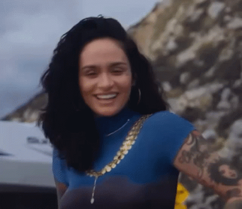 A Woman With A Tattoo On Her Arm Is Smiling While Wearing A Blue Turtleneck And A Gold Chain . GIF