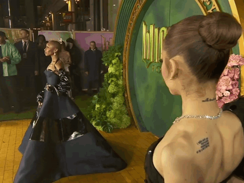 A Woman With A Tattoo On Her Back Stands In Front Of A Green Door That Says Wizard Of Oz GIF