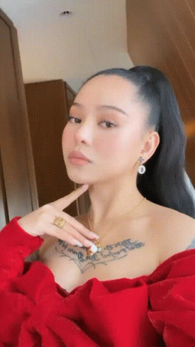 A Woman With A Tattoo On Her Chest Is Wearing A Red Top And Earrings GIF