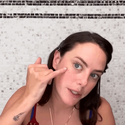A Woman With A Tattoo On Her Wrist Points To Her Face GIF