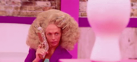A Woman With A Very Large Wig Is Holding A Gun In Front Of A Pink Balloon . GIF