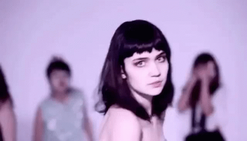 A Woman With Black Hair Is Standing In Front Of A Group Of People And Looking At The Camera . GIF