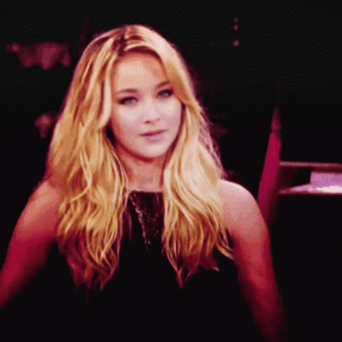 A Woman With Blonde Hair And A Black Dress Looks At The Camera GIF