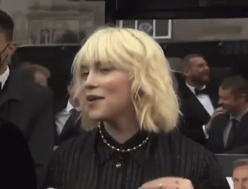 A Woman With Blonde Hair And A Black Shirt Is Standing In A Crowd . GIF