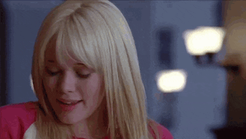 A Woman With Blonde Hair And Bangs Is Smiling While Wearing A Pink Shirt . GIF