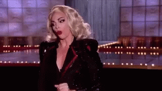 A Woman With Blonde Hair And Red Lips Is Standing On A Stage In A Black Jacket . GIF