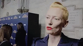 A Woman With Blonde Hair And Red Lipstick Is Standing In Front Of A Sign That Says ' Ladies Of The Race ' . GIF