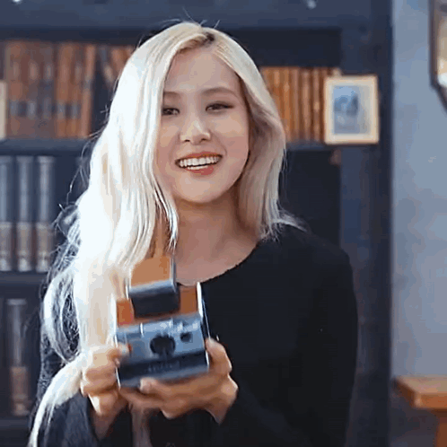 A Woman With Blonde Hair Is Holding A Camera In Her Hands And Smiling . GIF