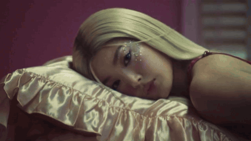A Woman With Blonde Hair Is Laying On A Bed With A Pink Pillow . GIF