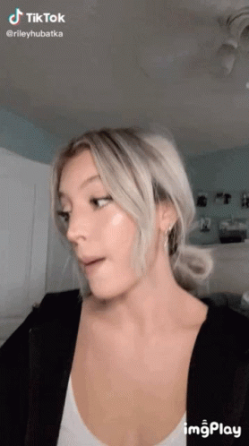 A Woman With Blonde Hair Is Making A Funny Face While Wearing A White Tank Top And A Black Jacket . GIF