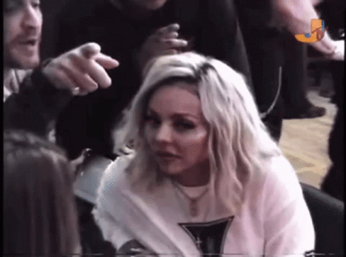 A Woman With Blonde Hair Is Sitting In A Crowd While A Man Points At Her . GIF