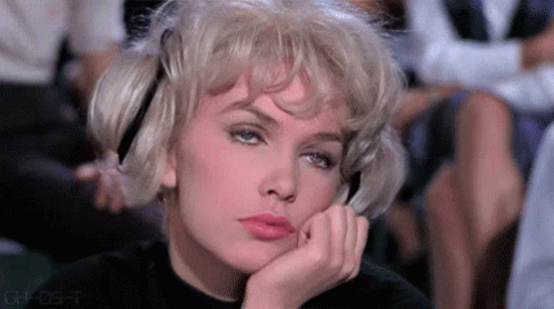 A Woman With Blonde Hair Is Sitting In A Theatre With Her Hand On Her Chin . GIF