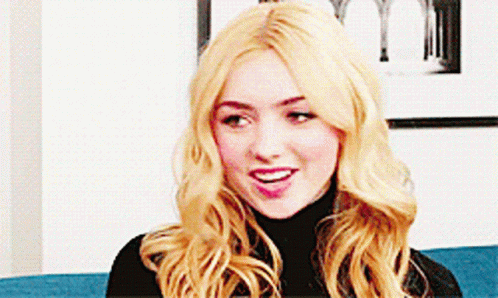 A Woman With Blonde Hair Is Sitting On A Blue Couch Wearing A Black Turtleneck . GIF
