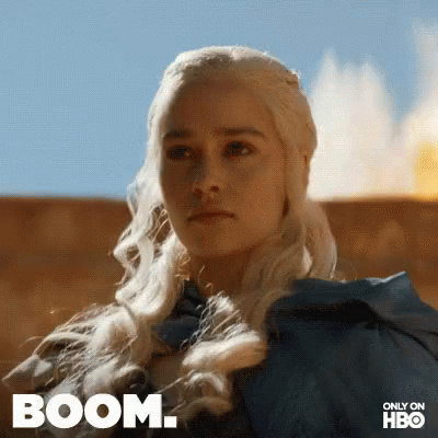 A Woman With Blonde Hair Is Standing In Front Of A Fire With The Words Boom Written On The Bottom . GIF