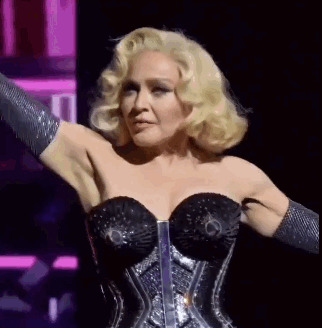 A Woman With Blonde Hair Is Wearing A Black Corset And Gloves On A Stage . GIF