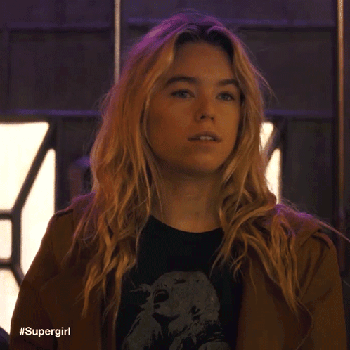A Woman With Blonde Hair Is Wearing A Brown Jacket And A Black Shirt With A Picture Of An Eagle On It . GIF