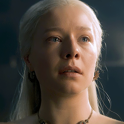 A Woman With Blonde Hair Wearing A Necklace And Earrings Looks At The Camera GIF