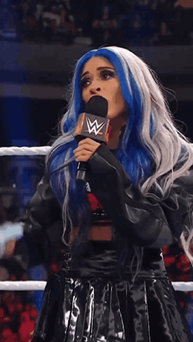 A Woman With Blue And White Hair Is Speaking Into A Microphone With A W Logo On It GIF