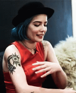 A Woman With Blue Hair And A Tattoo On Her Arm Is Wearing A Black Hat . GIF