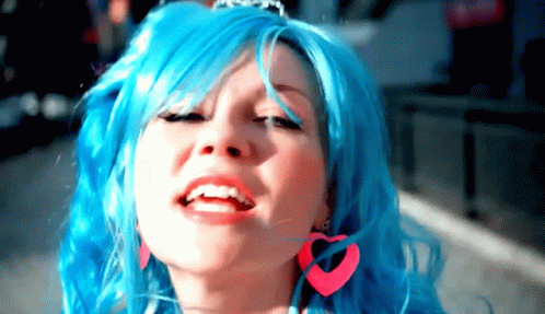A Woman With Blue Hair And Pink Heart Shaped Earrings Smiles GIF