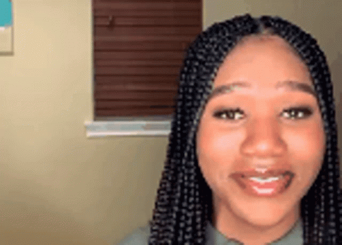 A Woman With Braids Is Smiling And Looking At The Camera In Front Of A Window . GIF