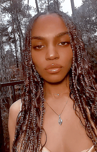 A Woman With Braids Is Wearing A Necklace With A Pendant GIF