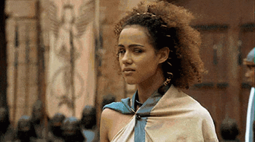 A Woman With Curly Hair And A Scarf Around Her Neck Is Standing In Front Of A Building . GIF