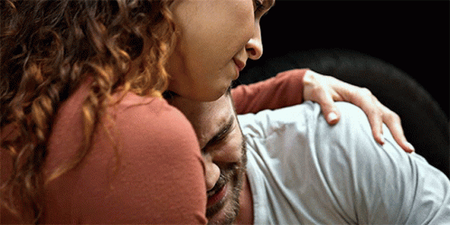 A Woman With Curly Hair Is Hugging A Man With His Eyes Closed GIF