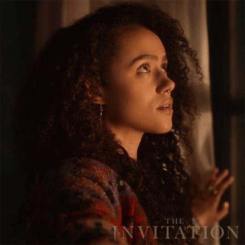 A Woman With Curly Hair Is Looking Out A Window With The Words The Invitation Behind Her GIF
