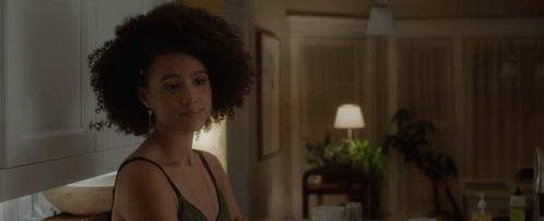 A Woman With Curly Hair Is Sitting At A Table In A Dark Room GIF