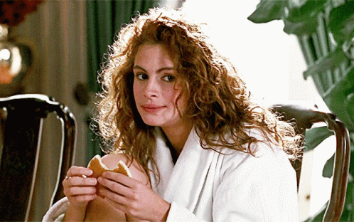 A Woman With Curly Hair Is Sitting In A Chair Holding A Sandwich GIF