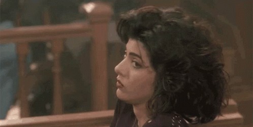 A Woman With Curly Hair Is Sitting On A Bench In Front Of A Wooden Railing . GIF