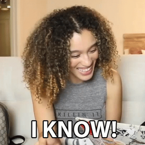 A Woman With Curly Hair Is Sitting On A Couch And Smiling While Holding A Magazine And Says I Know GIF
