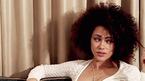 A Woman With Curly Hair Is Sitting On A Couch In Front Of A Curtain GIF