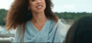 A Woman With Curly Hair Is Smiling And Looking At The Camera While Standing Next To Another Woman . GIF