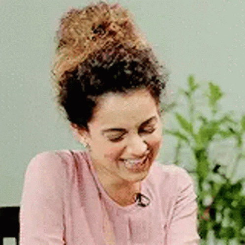 A Woman With Curly Hair Is Smiling While Wearing A Pink Shirt And Earrings . GIF