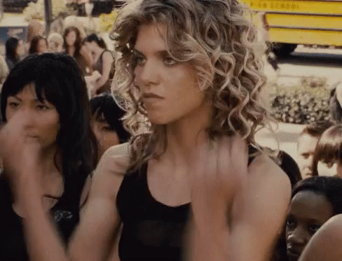 A Woman With Curly Hair Is Standing In Front Of A Crowd Of People And A Yellow School Bus GIF
