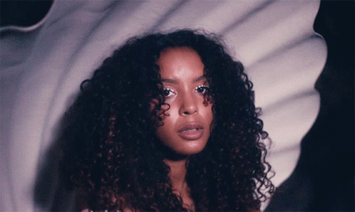 A Woman With Curly Hair Is Standing In Front Of A White Background GIF